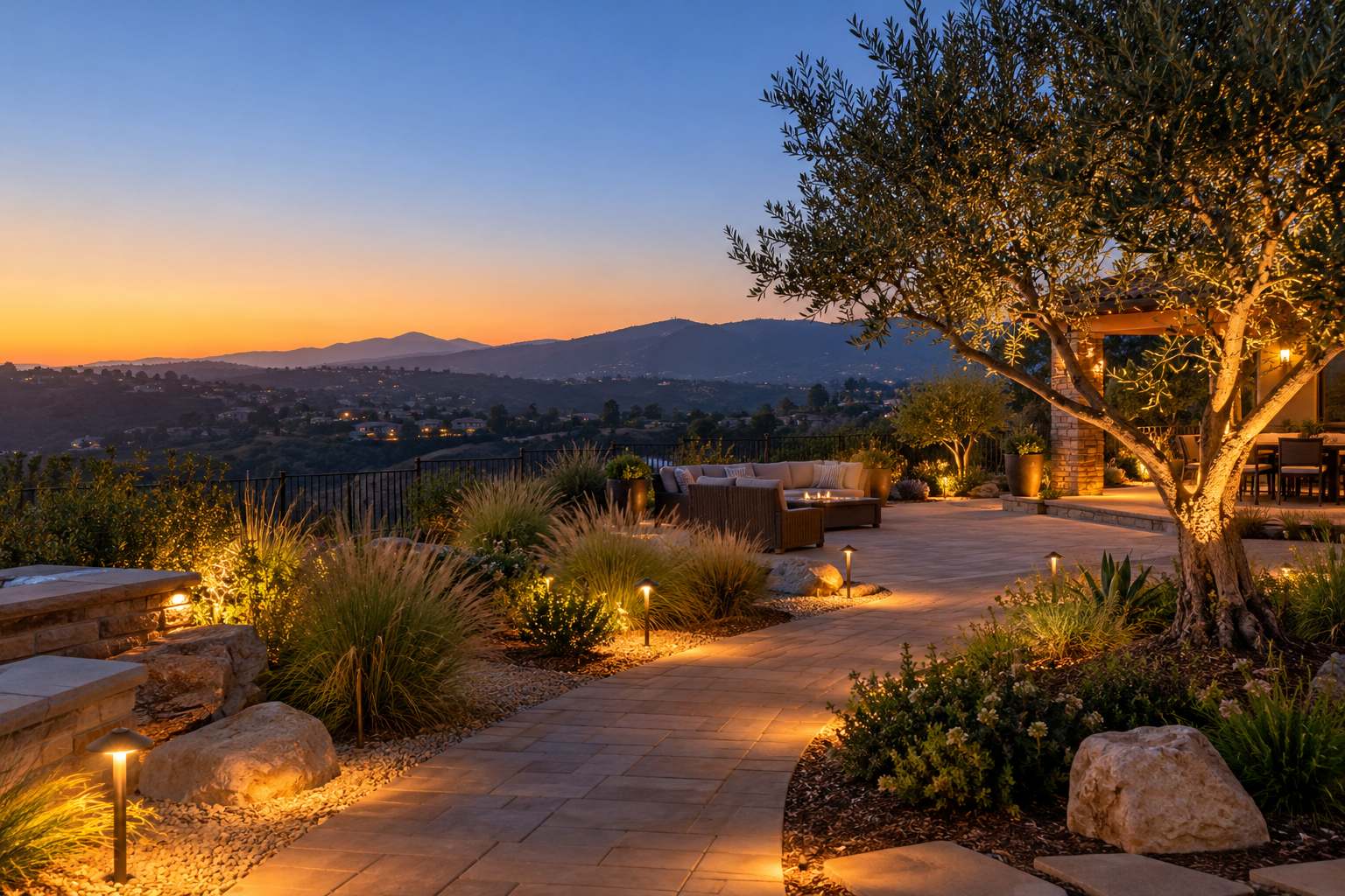 Backyard at dusk with lit paver paths, an uplit olive tree and seating beyond